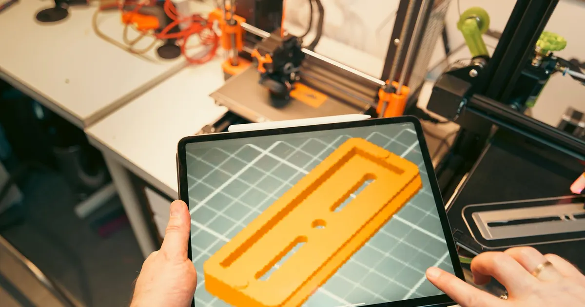 Tablet showing a 3D model of a flat part with slots, held in a workshop during a CNC prototype design iteration review